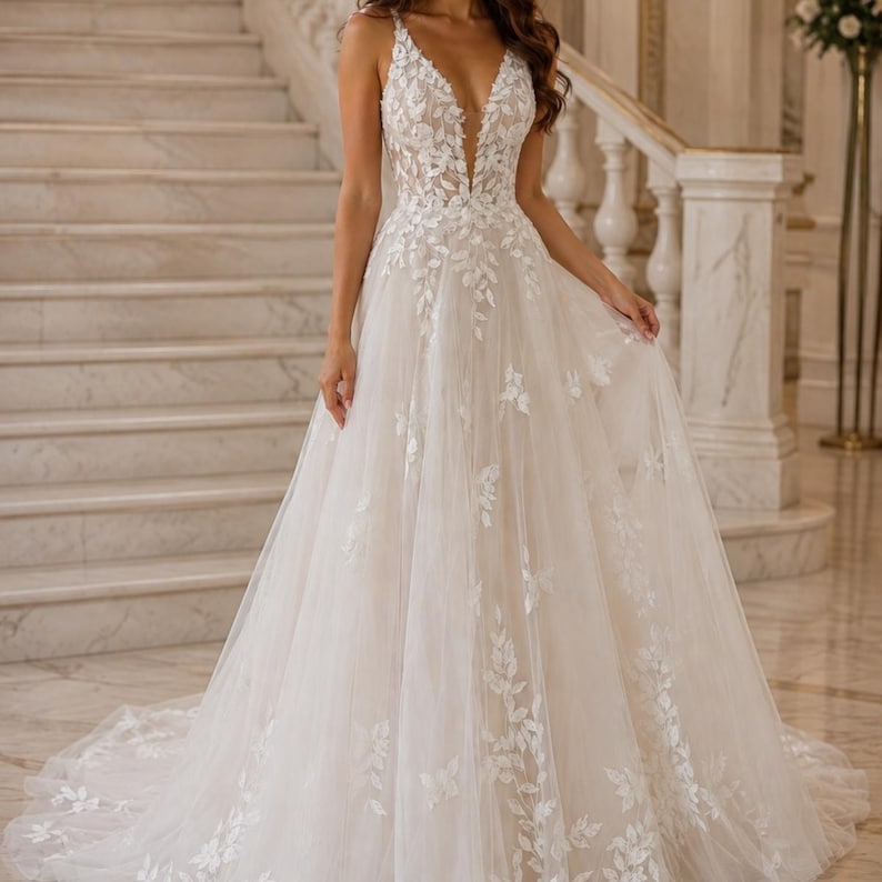 May include: A floor-length A-line gown in an off-white shade featuring a deep V-neckline and thin straps. The bodice and skirt are adorned with intricate white leaf-shaped lace appliqu&eacute;s that cascade down the layered tulle fabric to the hem.