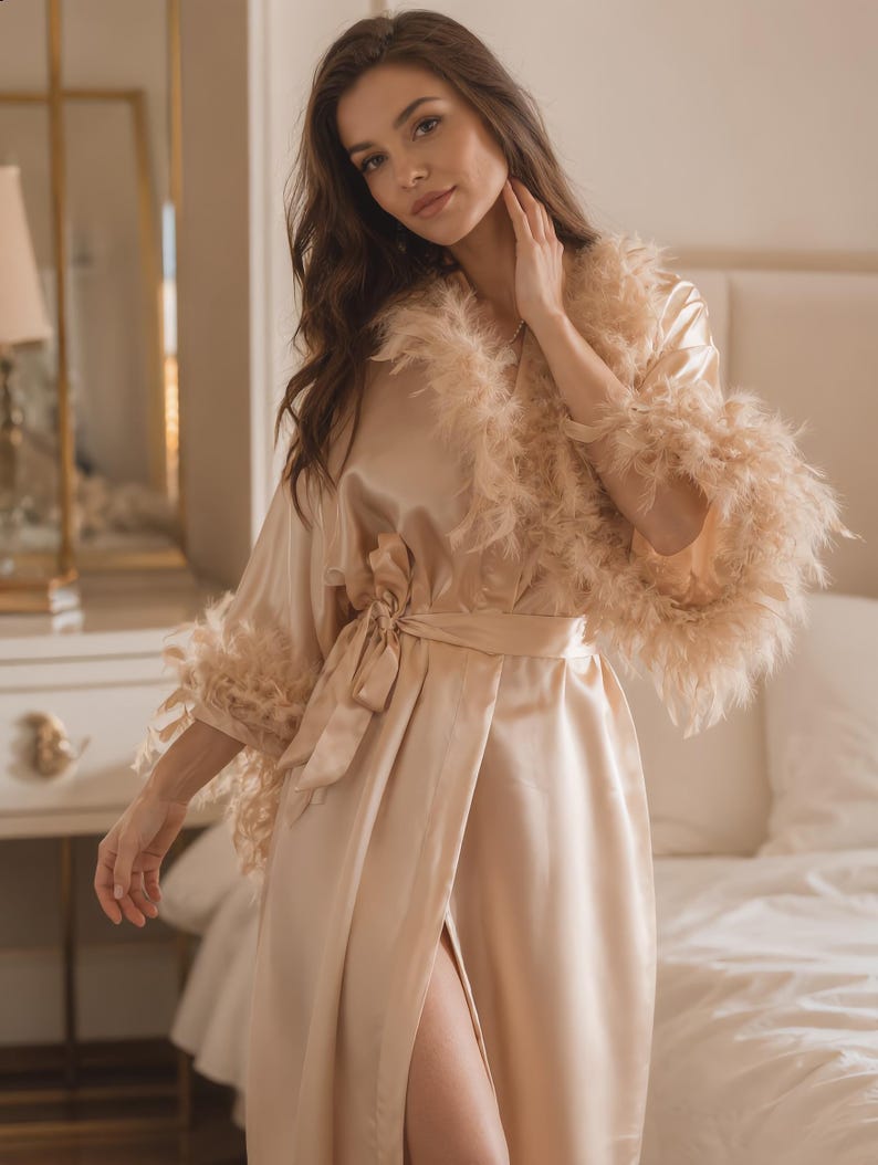 May include: A light peach-colored satin robe with feather trim on the sleeves and collar. The robe has a matching tie belt and a thigh-high slit. The background includes a bed and a vanity.