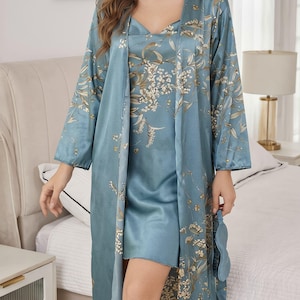 May include: A three-piece sleepwear set in a light blue hue, featuring a satin robe, a slip dress, and an eye mask. The set is adorned with a floral pattern in shades of white and gold. The robe has long sleeves and a flowing design.