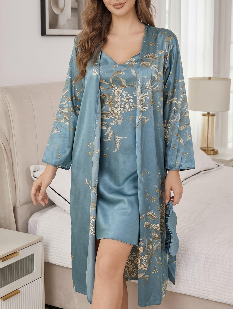 May include: A three-piece sleepwear set in a light blue hue, featuring a satin robe, a slip dress, and an eye mask. The set is adorned with a floral pattern in shades of white and gold. The robe has long sleeves and a flowing design.