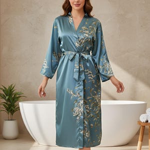May include: A teal satin robe with a floral pattern and a matching belt. The robe has long sleeves and a V-neck. The floral design features white and gold accents. The robe is knee-length and appears to be made of a silky material.