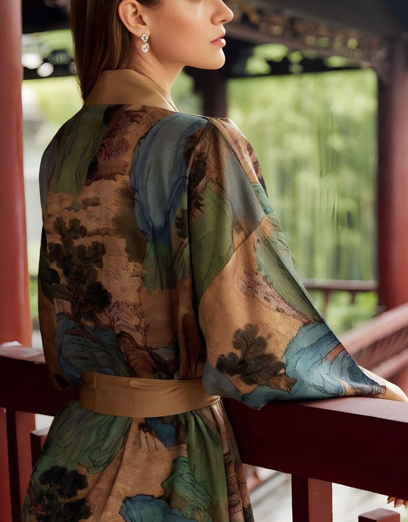 K&ouml;nnte beinhalten: Seidenkimono mit Landschaftsmuster in Gr&uuml;n-, Blau- und Braunt&ouml;nen. Der Kimono hat einen breiten, beigefarbenen G&uuml;rtel und Besatz. Die &Auml;rmel sind lang und flie&szlig;end. Die Person steht auf einem roten Holzbalon.