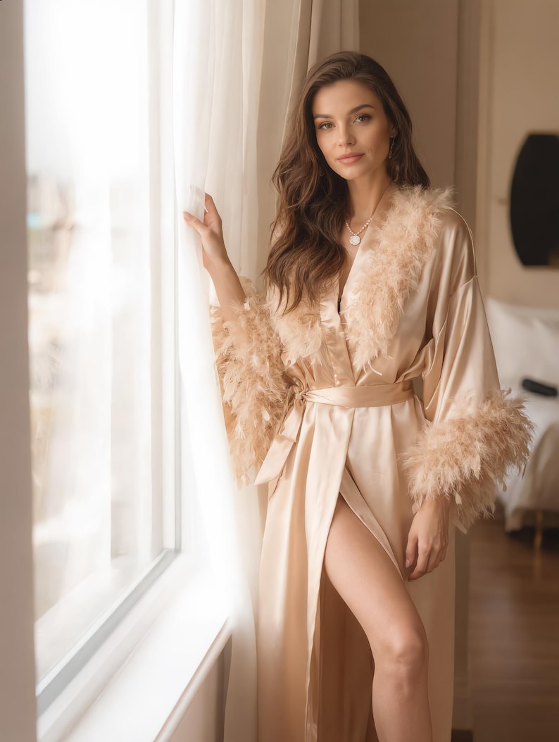 May include: A champagne-colored satin robe with feather trim on the collar and cuffs. The robe is belted at the waist and has a high slit. The model is standing near a window.
