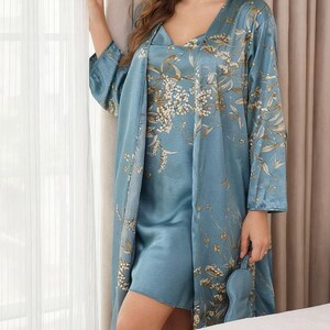 May include: A light blue satin pajama set with a floral pattern. The set includes a short-sleeved top, a long-sleeved robe, and an eye mask. The floral design features white and gold accents. The robe is knee-length.