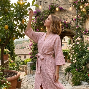 May include: A pink leopard print robe with a matching belt. The robe has wide sleeves and a slit up the front. The image is taken outdoors with a lemon tree and a stone building in the background.