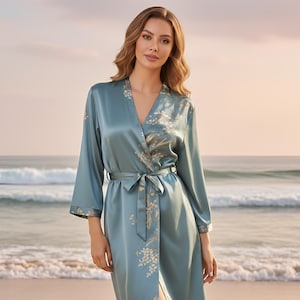 May include: A light blue satin robe with floral embroidery. The robe features a V-neck, long sleeves, and a matching belt. The floral design is in a cream colour. The robe is knee-length and is worn on a beach.