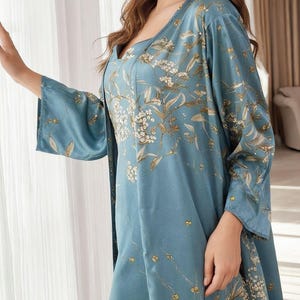 May include: A light blue satin sleepwear set featuring a floral pattern in white and gold. The set includes a square-neck slip dress and a matching robe with wide sleeves. The fabric has a subtle sheen, and the floral design adds a touch of elegance.