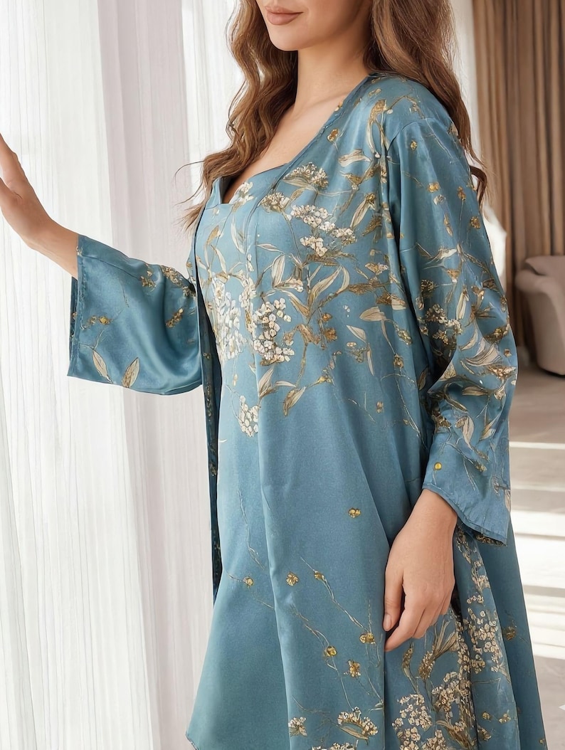 May include: A light blue satin sleepwear set featuring a floral pattern in white and gold. The set includes a square-neck slip dress and a matching robe with wide sleeves. The fabric has a subtle sheen, and the floral design adds a touch of elegance.