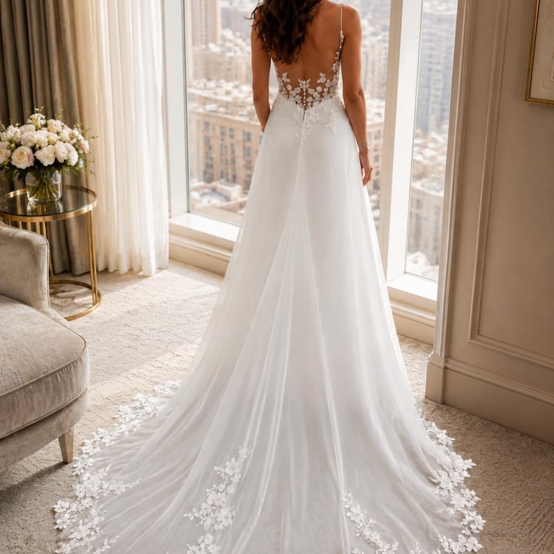 May include: A floor-length white wedding dress featuring a sheer back with intricate floral lace embroidery, thin straps, and a flowing skirt with a long train adorned with matching lace appliqu&eacute;s along the hemline and a fitted bodice.