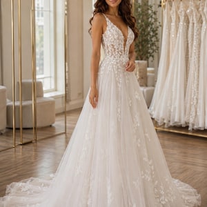 May include: A floor-length ivory wedding dress featuring a deep V-neckline and spaghetti straps. The bodice and skirt are adorned with intricate white floral lace appliqu&eacute;s that cascade down the layered tulle fabric, ending in a long train.