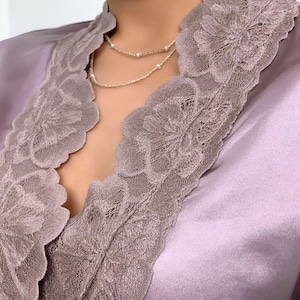 Puede incluir: Primer plano de una bata de sat&eacute;n color lavanda con intrincados adornos de encaje. Un delicado collar de plata con peque&ntilde;as perlas descansa sobre el pecho. El encaje de la bata presenta un dise&ntilde;o floral.