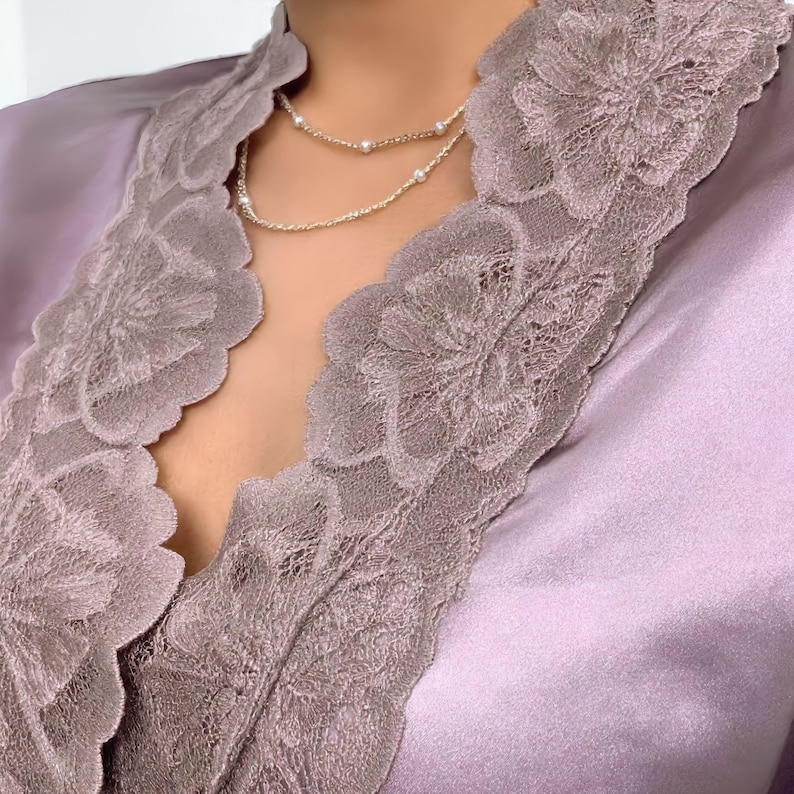 Puede incluir: Primer plano de una bata de sat&eacute;n color lavanda con intrincados adornos de encaje. Un delicado collar de plata con peque&ntilde;as perlas descansa sobre el pecho. El encaje de la bata presenta un dise&ntilde;o floral.