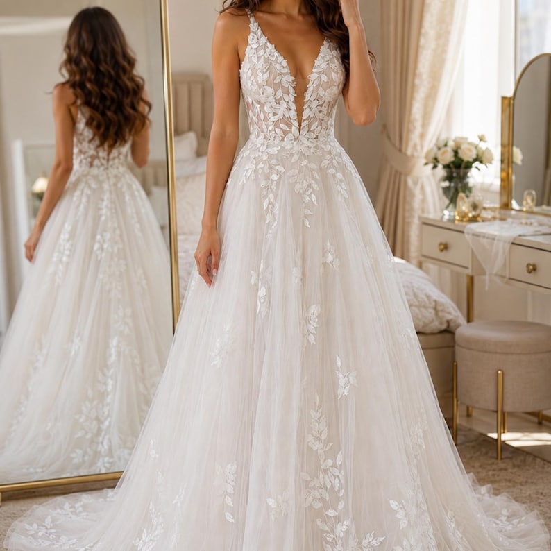 May include: A floor-length white wedding dress featuring a deep V-neckline and thin straps. The bodice and skirt are adorned with intricate white leaf-patterned lace appliqu&eacute;s that cascade down the tulle fabric, creating a full, flowing silhouette.