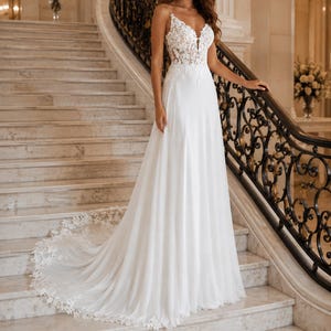 May include: A floor-length white gown featuring a sleeveless bodice with intricate floral lace appliques, a deep V-neckline, and a flowing skirt that extends into a long train with a scalloped lace hemline detail along the bottom edge.