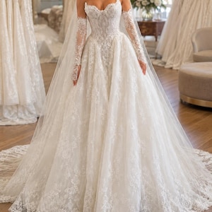 May include: An ivory strapless ballgown wedding dress featuring a sweetheart neckline, intricate lace embroidery throughout the bodice and skirt, a full tulle skirt with a scalloped lace hem, and matching sheer lace long sleeves with finger loops.
