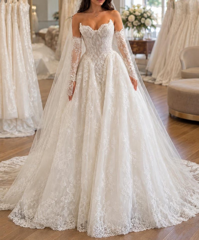 May include: An ivory strapless ballgown wedding dress featuring a sweetheart neckline, intricate lace embroidery throughout the bodice and skirt, a full tulle skirt with a scalloped lace hem, and matching sheer lace long sleeves with finger loops.