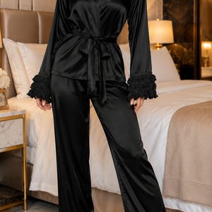May include: A black satin pyjama set consisting of a long-sleeved button-down top with a tie waist and matching wide-leg trousers. The garment features black lace detailing on the shoulders and tiered ruffled cuffs on the sleeves and trouser hems.
