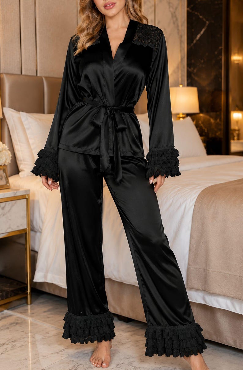 May include: A black satin pyjama set consisting of a long-sleeved button-down top with a tie waist and matching wide-leg trousers. The garment features black lace detailing on the shoulders and tiered ruffled cuffs on the sleeves and trouser hems.
