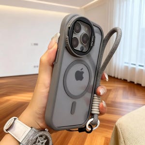 May include: Gray phone case with a clear camera lens protector and a gray lanyard. The case has a circular Apple logo and a magnetic ring on the back. The lanyard is attached to the case with a white and gray cord.