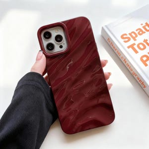 Pode incluir: Capa de telefone bordô com uma superfície brilhante e ondulada. A capa tem uma borda vermelha escura ao redor das lentes da câmera. Um livro com o título "Spati Tot P" é visível no fundo.
