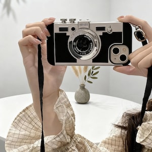 May include: A black and silver phone case designed to resemble a vintage camera. The case features a lens, buttons, and a strap for carrying. The phone case has the text "02-100" on the top.