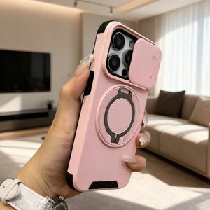 May include: A pink phone case with a black border and a camera cover. The case has a ring holder on the back and is designed to protect the phone's camera lenses. The case is held in a person's hand.