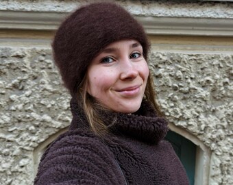Könnte beinhalten: Eine Person trägt einen dunkelbraunen, flauschigen Hut und einen passenden Teddybär-Mantel. Der Mantel hat einen hohen Rollkragen. Ein schwarzer Riemen einer Tasche ist über der Schulter zu sehen. Der Hintergrund ist ein strukturiertes, hellfarbiges Gebäude.