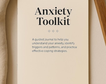Op de afbeelding: Een crèmekleurig tijdschrift met de titel "Anxiety Toolkit" met zwarte tekst. De beschrijving van het tijdschrift luidt: "Een geleid tijdschrift om u te helpen uw angst te begrijpen, triggers en patronen te identificeren en effectieve copingstrategieën te oefenen."