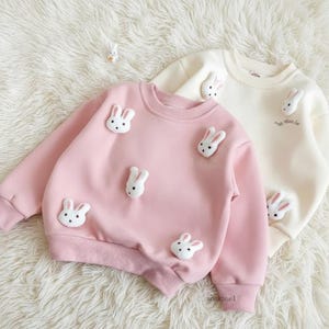 May include: Two children's sweatshirts, one pink and one cream, adorned with white bunny appliqués. The pink sweatshirt has multiple bunny faces, while the cream one has a few. Both have ribbed cuffs and a crew neck, perfect for a cozy look.