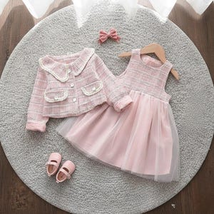 May include: A pink and white plaid jacket with a matching dress and shoes. The jacket has a white collar and pockets. The dress has a tulle skirt. A pink bow and matching shoes complete the outfit. The outfit is for a child.