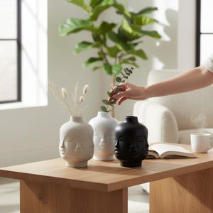Op de afbeelding: Drie keramische gezichtsvazen in wit en zwart staan op een houten tafel. De vazen hebben een uniek ontwerp met meerdere gezichten. Eén vaas bevat droogbloemen, terwijl een andere wordt gestyled met groen. De vazen zijn een decoratief woonaccessoire.