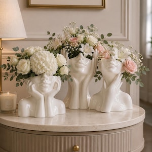 May include: Three white ceramic face vases filled with flowers, including roses and hydrangeas, sit on a round table. The vases have different poses, with hands supporting the face or holding the flowers. A lamp and a candle are in the background.