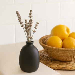 Puede incluir: Un pequeño jarrón negro mate con ramitas de lavanda seca. Una cesta de mimbre llena de limones amarillos brillantes se encuentra cerca sobre un salvamanteles de tonos neutros. La escena se desarrolla contra una pared de azulejos blancos.