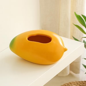 Pode incluir: Um vaso decorativo de cerâmica em forma de manga. O vaso é principalmente amarelo com uma ponta verde e um interior castanho. Possui uma grande abertura oval na parte superior. O vaso está sobre uma prateleira branca.