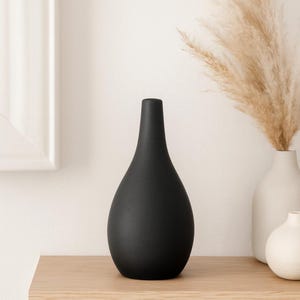 May include: A black, teardrop-shaped vase with a narrow neck. The vase is made of a matte material and sits on a light wooden surface. A white vase and dried pampas grass are visible in the background, creating a minimalist aesthetic.