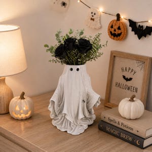 May include: A white ceramic ghost-shaped vase featuring two small black eyes, filled with a bouquet of dark black roses and green foliage. The vase has a draped, fabric-like texture and a small spider detail on the side, creating a spooky aesthetic.