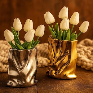 May include: Two decorative vases, one silver and one gold, each holding white tulips with green stems and leaves. The vases have a unique, crumpled paper bag design. The background features a wooden surface and a soft, textured blanket.