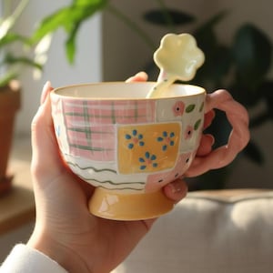 Pode incluir: Uma caneca de cerâmica com um design de patchwork com xadrez rosa, quadrados amarelos com flores azuis e padrões florais rosa. A caneca tem uma alça rosa torcida e uma colher a condizer com uma cabeça em forma de flor. A caneca está numa mão.