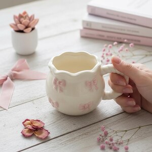 Puede incluir: Una taza de cerámica blanca con borde festoneado y asa. La taza está adornada con lazos rosas en relieve. Una pequeña suculenta en una maceta blanca y una cinta rosa están en el fondo.