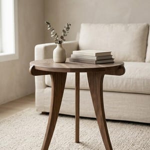 Könnte beinhalten: Ein runder Beistelltisch aus dunklem Holz mit drei geschwungenen Beinen. Auf dem Tisch stehen eine kleine Vase mit Grünzeug und ein Stapel Bücher. Der Tisch hat einen Durchmesser von ca. 61 cm.