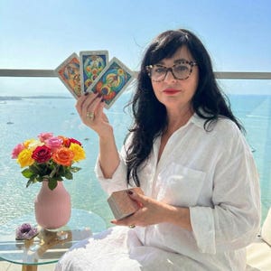 May include: A woman holding tarot cards, with a deck in her lap. A vase of colorful roses sits on a table. The background shows a body of water and a clear sky.