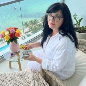May include: A woman seated on a balcony with a view of the ocean, holding tarot cards. A small glass table holds a vase of colorful roses and a purple crystal. The woman wears a white shirt and glasses.