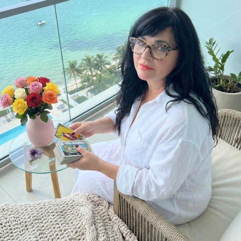 May include: A woman seated on a balcony with a view of the ocean, holding tarot cards. A small glass table holds a vase of colorful roses and a purple crystal. The woman wears a white shirt and glasses.