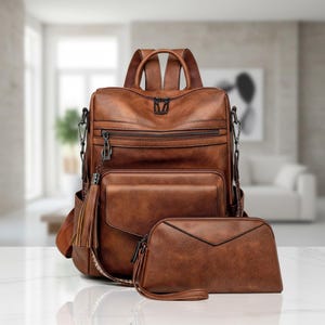 Puede incluir: Mochila de cuero sintético marrón con un clutch a juego. La mochila tiene múltiples compartimentos con cremallera, un bolsillo frontal y una borla decorativa. El clutch tiene una correa de muñeca. El conjunto está diseñado para el uso diario.