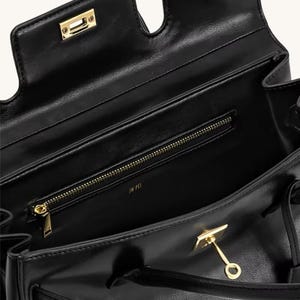 Op de afbeelding: Zwarte leren handtas met goudkleurige hardware. De tas heeft een handvat aan de bovenkant, een binnenvak met ritssluiting en een klepsluiting met een gouden sluiting. De binnenkant is gevoerd met een glad materiaal. De merknaam "JW PEI" is zichtbaar in de tas.