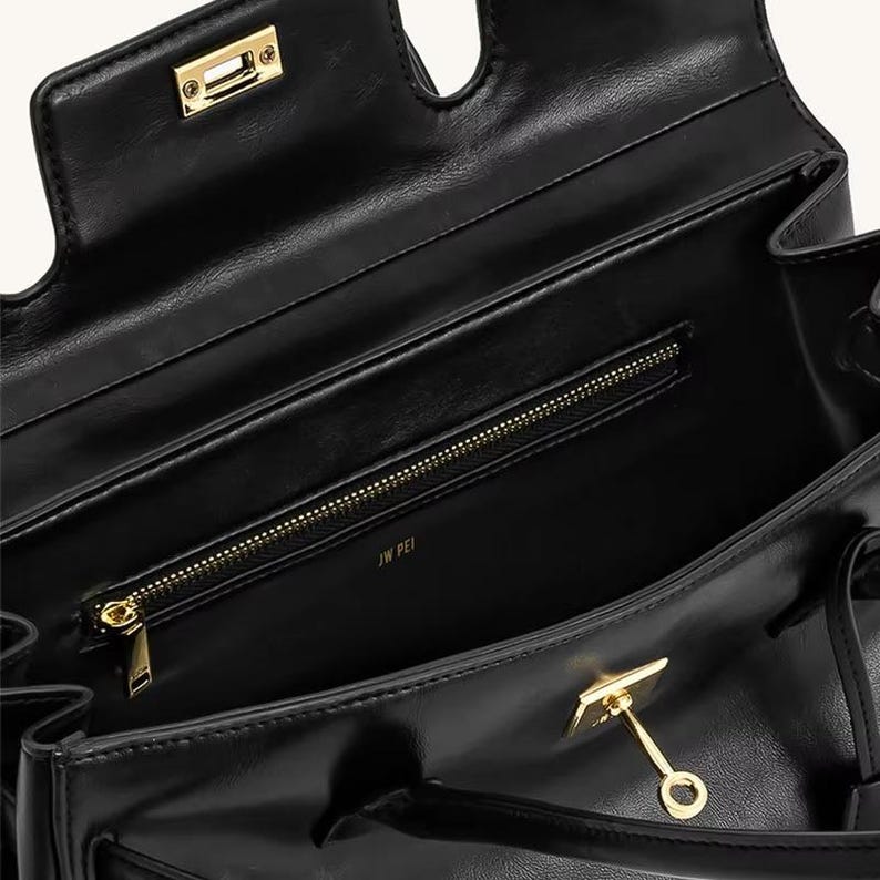 Op de afbeelding: Zwarte leren handtas met goudkleurige hardware. De tas heeft een handvat aan de bovenkant, een binnenvak met ritssluiting en een klepsluiting met een gouden sluiting. De binnenkant is gevoerd met een glad materiaal. De merknaam "JW PEI" is zichtbaar in de tas.