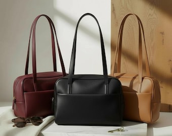 May include: Three leather handbags in burgundy, black, and tan. Each bag has a rectangular shape, a top handle, and a front pocket. Sunglasses and a key rest on a white surface in front of the bags.