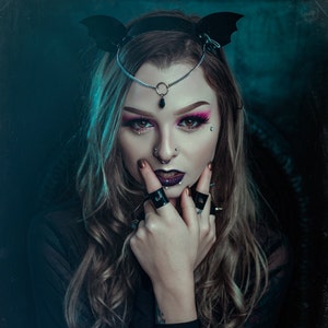 May include: A woman with dark hair and dark makeup is wearing a black bat headband, a silver chain forehead piece with a black pendant, and multiple rings on her fingers. She is looking directly at the camera.