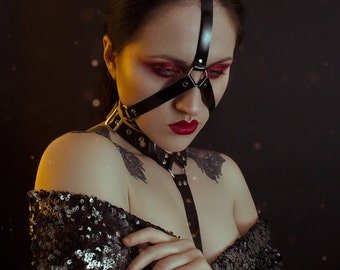 May include: A woman with dark hair and red lipstick wears a black leather harness with a strap across her eyes. She is wearing a sequined top and has a dark background with a subtle sparkle.