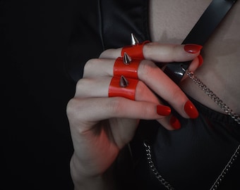 May include: Red finger armor with silver spikes. The image shows a close-up of a hand wearing the armor, with a black background. The nails are painted red. The logo "KATZ LITTLE FACTORY" is in the bottom left corner.